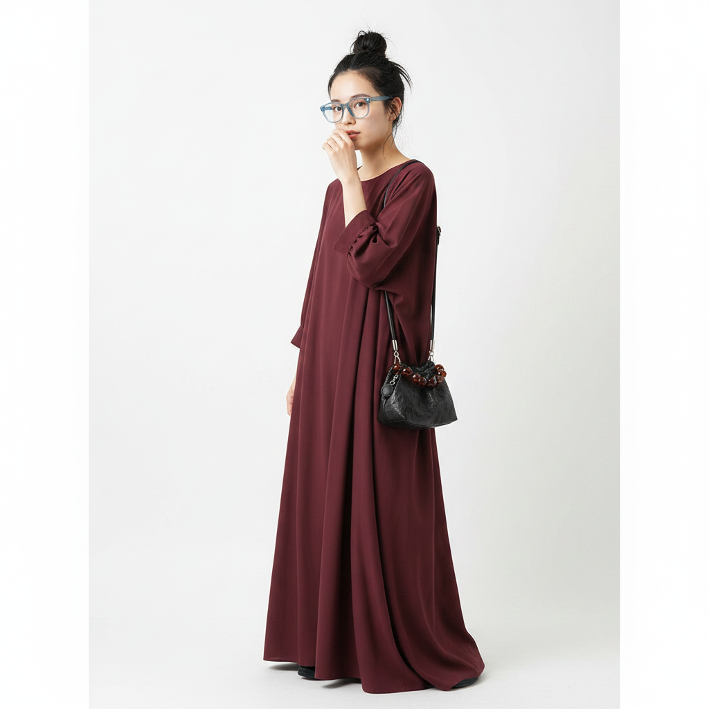 A woman wearing a long, loose-fitting burgundy maxi dress with gathered sleeves and side ties, styled with a black shoulder bag, standing against a plain light gray background.