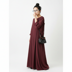 A woman wearing a long, loose-fitting burgundy maxi dress with gathered sleeves and side ties, styled with a black shoulder bag, standing against a plain light gray background.