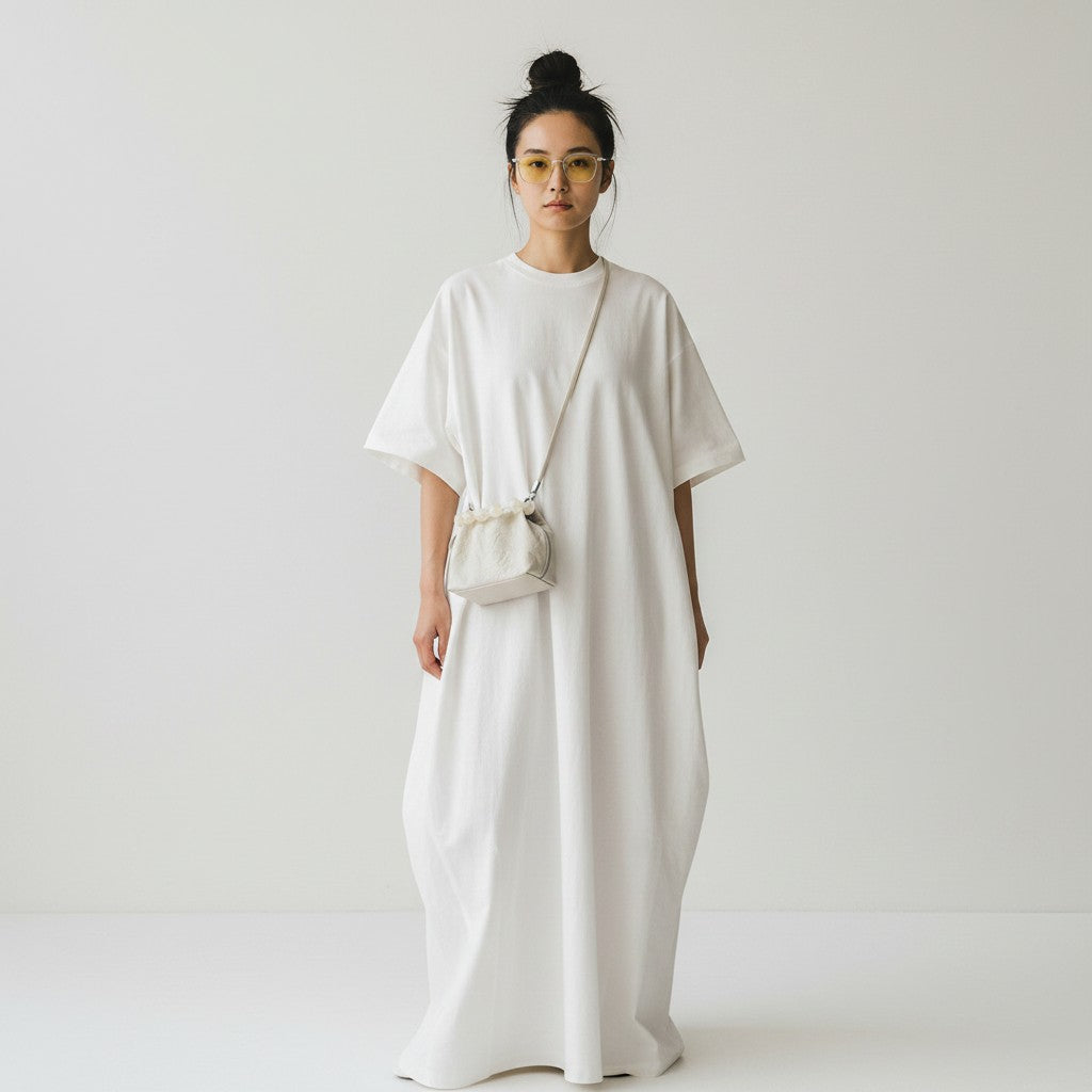 A woman wearing a loose, oversized white short-sleeve maxi dress and a small white crossbody bag, standing against a plain light gray studio background.