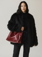 A model wearing a black faux fur coat, black turtleneck, and black jeans, carrying a large burgundy leather shoulder bag against a light gray studio background.