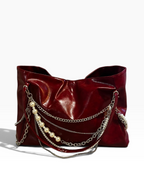 A glossy deep red leather shoulder bag featuring layered silver chains and a strand of white faux pearls draped over the front, set against a plain white background.