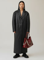 A model stands against a light gray studio background wearing a long black leather-like coat, black boots, and holding a burgundy tote bag with silver chain details.