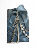 A close-up of a blue leather bag featuring multiple hanging chain straps, including a pearl bead strand and a silver-toned beaded chain, against a plain white background.