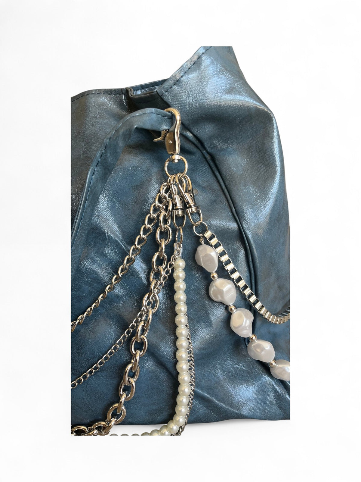 A close-up of a blue leather bag featuring multiple hanging chain straps, including a pearl bead strand and a silver-toned beaded chain, against a plain white background.