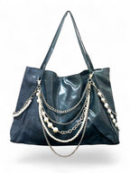 A black faux-leather hobo tote bag featuring multiple draped silver chains and strands of white faux pearls on the front, set against a plain white background.