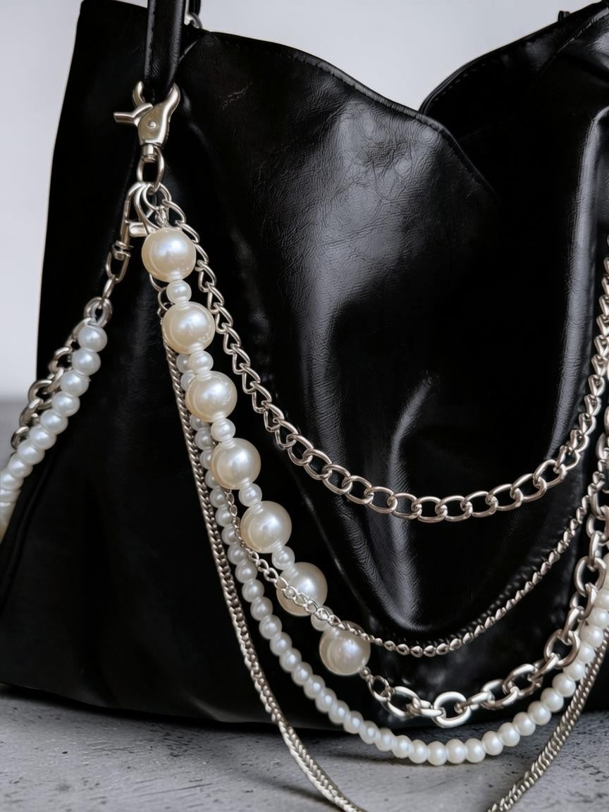 A close-up of a black leather shoulder bag featuring multiple draped chain straps and pearl bead accents in silver-tone hardware.