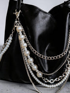 A close-up of a black leather shoulder bag featuring multiple draped chain straps and pearl bead accents in silver-tone hardware.